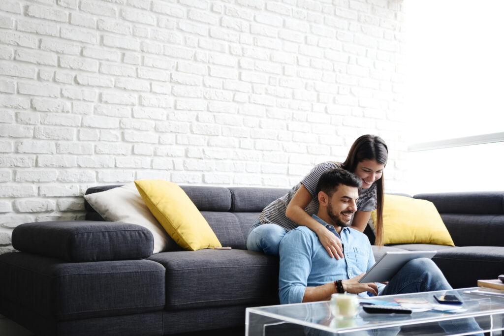 Young hispanic couple sitting on couch at home, using a tablet PC for Internet and social media. The girl is giving a massage to her boyfriend. Copy space